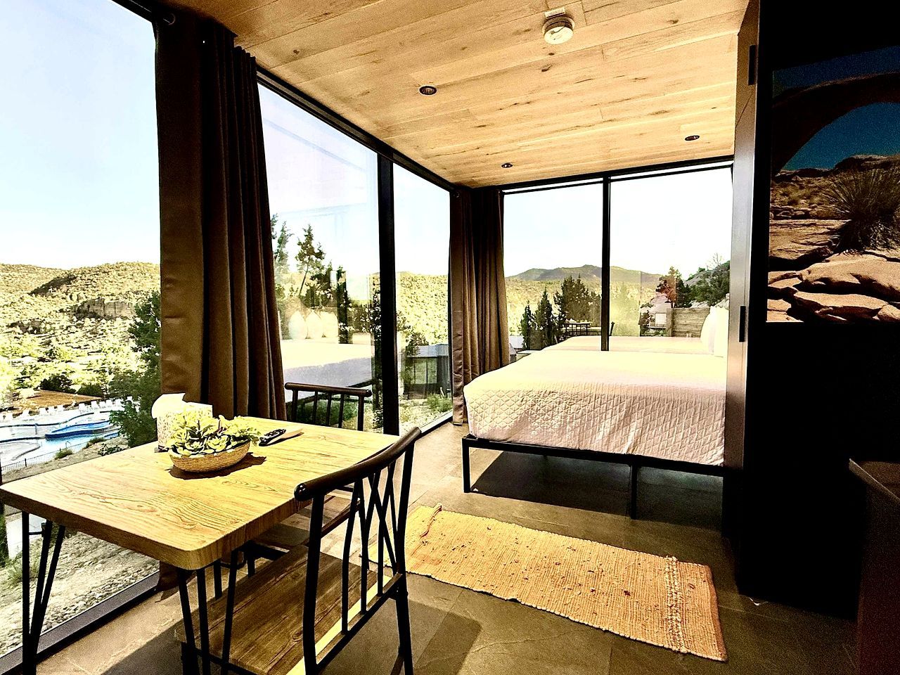 A bedroom with a large bed, a wooden table with a basket, and a view of a landscape through large windows.