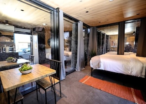 A modern bedroom with a large bed, a dining area with a table and chairs, and a view through a glass door. - Mirror Tiny House with Equipped Interior and Fire Pit in Orderville, Utah (Orderville)