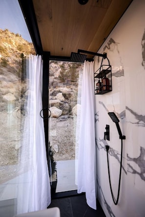 A modern bathroom with a shower, white curtains, and a view of rocky terrain. - Mirror Tiny House with Equipped Interior and Fire Pit in Orderville, Utah (Orderville)