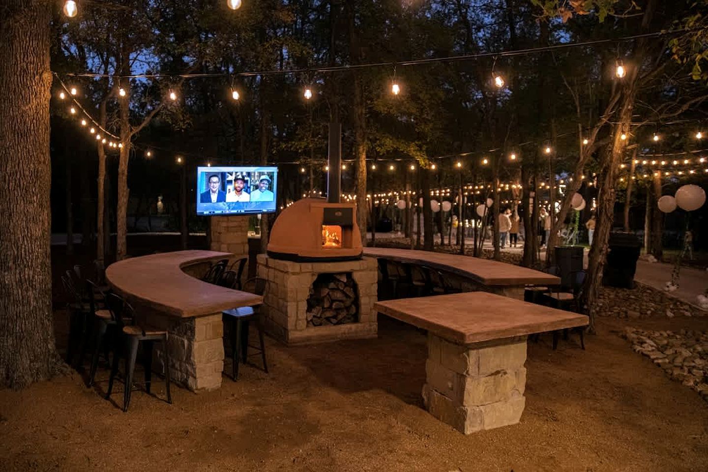 An outdoor patio with stone tables and chairs, a stone fireplace, and string lights.