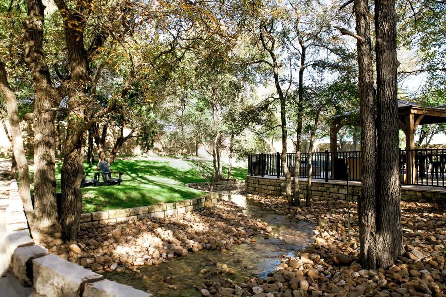 A park with trees, a stone pathway, a small stream, and a gazebo.