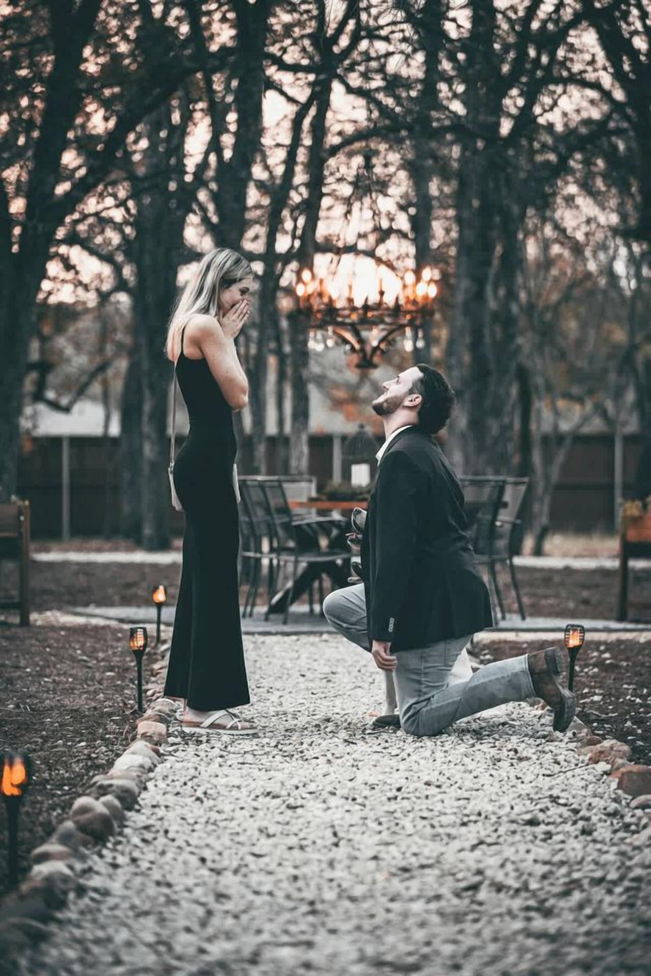 A man kneeling on a gravel path, proposing to a woman standing nearby in a garden setting with trees and a table in the background.