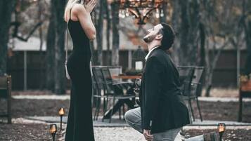 A man kneeling on a gravel path, proposing to a woman standing nearby in a garden setting with trees and a table in the background.