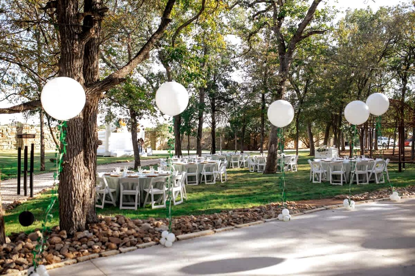 An outdoor event space with round tables and chairs, surrounded by trees and a stone wall.