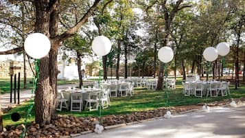 An outdoor event space with round tables and chairs, surrounded by trees and a stone wall.