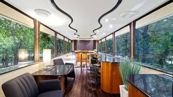 A modern interior with a long table, chairs, a bar area, and large windows offering a view of the outdoors.