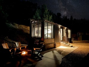 A glass-enclosed structure with a fire pit and seating area, surrounded by a gravel path and trees. - Unique Tiny House with Outdoor BBQ in Orderville, Utah (Orderville)