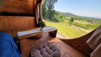 A wooden interior with a bed, a bench, and a cushioned seating area. A window offers a view of a green landscape.
