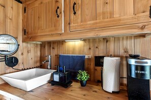 Innenansicht einer Holzhütte mit einer weißen Badewanne, einem Waschbecken, einem Spiegel, einem Handtuch, einer Pflanze und einer Keurig-Kaffeemaschine.