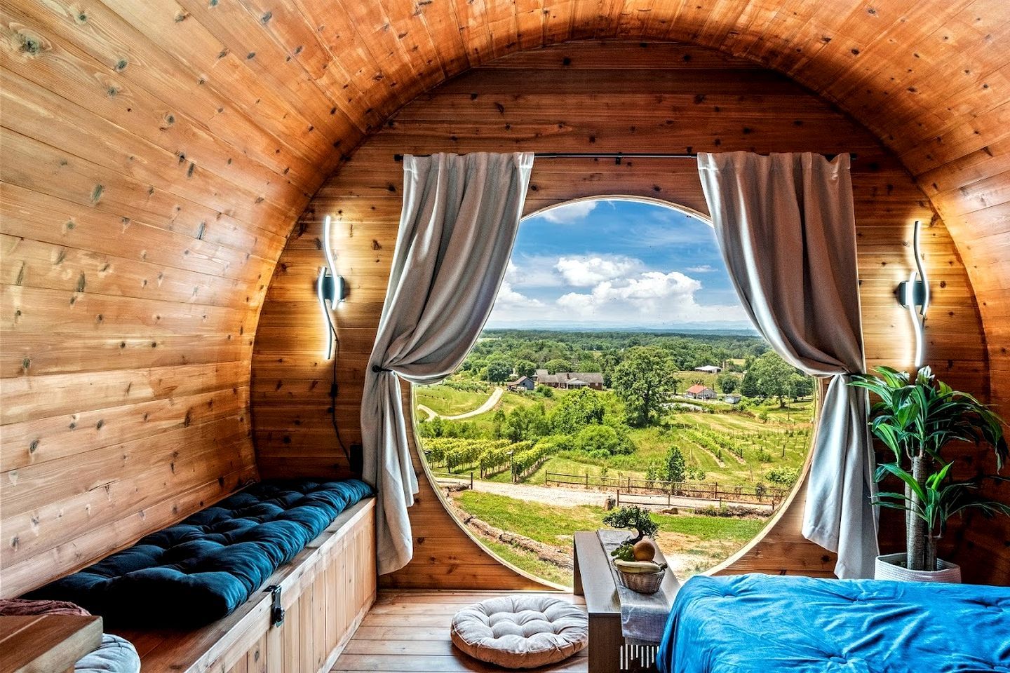 Un intérieur chaleureux, avec des murs en bois et un plafond voûté, comprenant un lit, un banc et offrant une vue sur un paysage luxuriant.