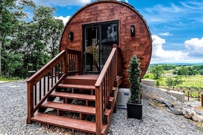 A wooden cabin with a staircase leading to a door, surrounded by gravel and greenery.