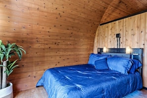 Ein Schlafzimmer mit Holzwänden, einem Bett mit blauer Bettwäsche, einem Nachttisch, einer Lampe und einem Topf mit Pflanze.