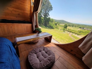 Ein Holzinnenraum mit einem Bett, einer Bank und einer gepolsterten Sitzecke. Ein Fenster bietet Blick auf eine grüne Landschaft.