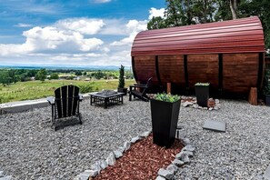 Et rustikt udendørs opholdsområde med Adirondack-stole og bålsted, omgivet af grus og træflis, med en stor træfadskonstruktion i baggrunden.