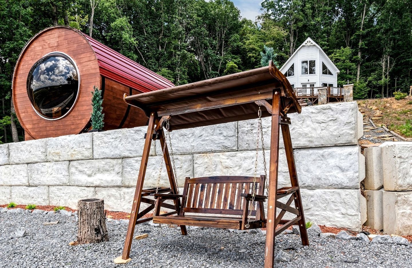 A rustic wooden swing set in a natural setting with a stone wall and a large wooden barrel in the background.