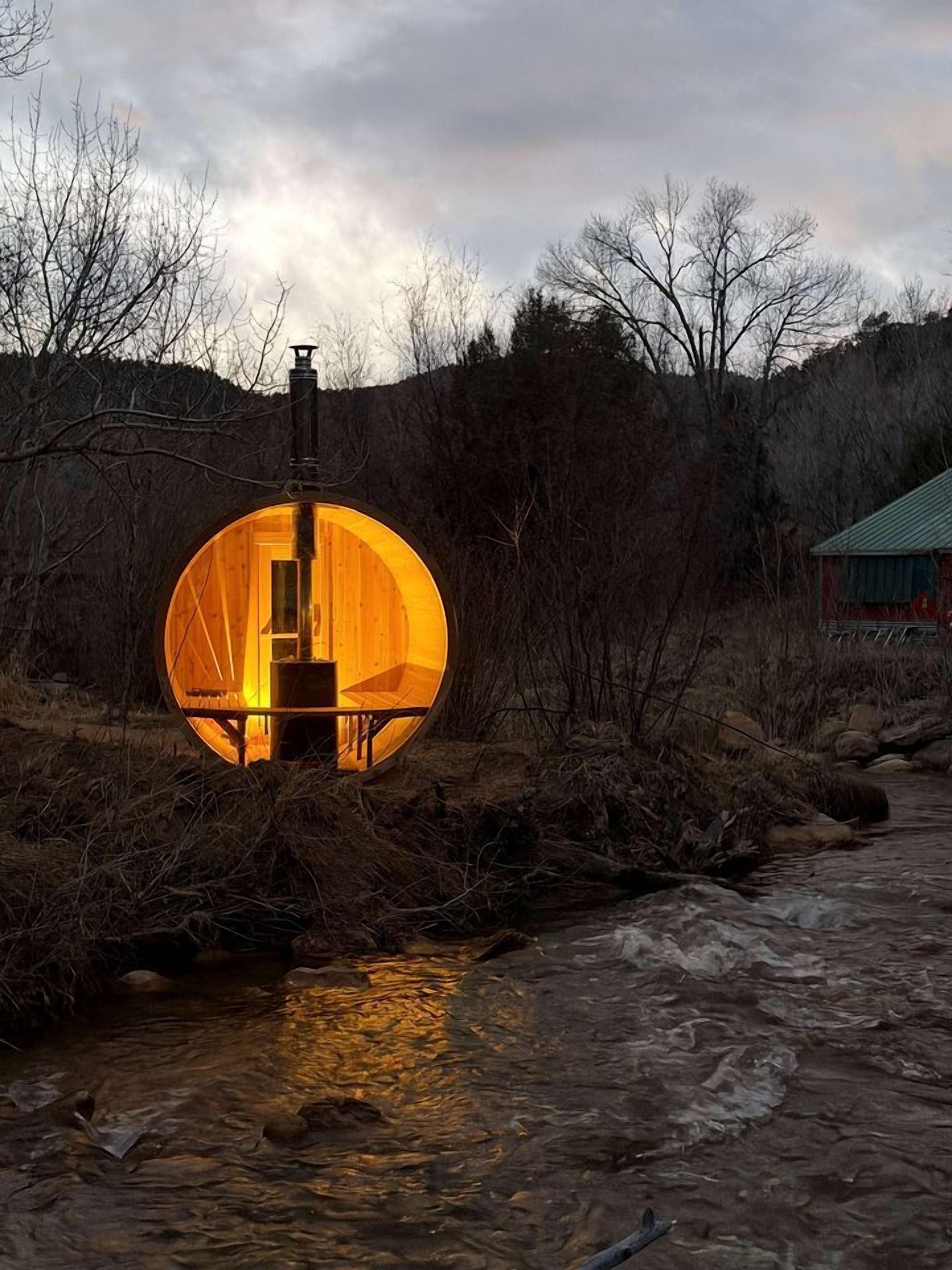 Une structure circulaire dotée d’un intérieur en bois, située au bord d’un ruisseau, entourée d’arbres et d’un bâtiment visible au loin.