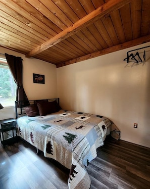 Ein Schlafzimmer mit Holzboden, einem Bett mit einem gemusterten Quilt, einem Nachttisch, einem Fenster mit Vorhängen und einer wandmontierten Leuchte.