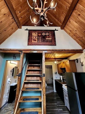 A wooden staircase leading to a kitchen with a chandelier and a decorative wall hanging.