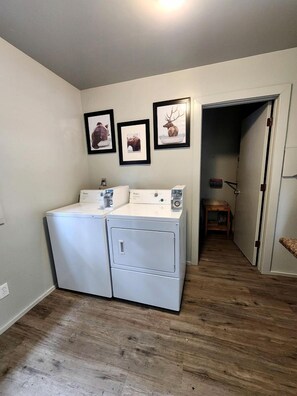 A laundry room with two washing machines and a dryer, a wooden floor, and a doorway leading to another room. - Fantastic Cabin Next to Wonderful Hiking Trails in Seward, Alaska (Seward)
