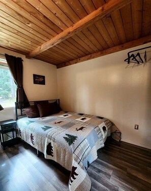 A bedroom with a wooden ceiling, a bed with a patterned quilt, a nightstand, a window with curtains, and a wall-mounted light fixture. - Fantastic Cabin Next to Wonderful Hiking Trails in Seward, Alaska (Seward)