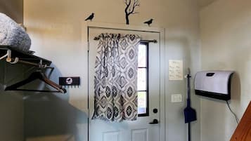 Une pièce avec une porte blanche, des rideaux à motifs, un plancher en bois, un porte-manteau suspendu, une boîte aux lettres fixée au mur et un balai.