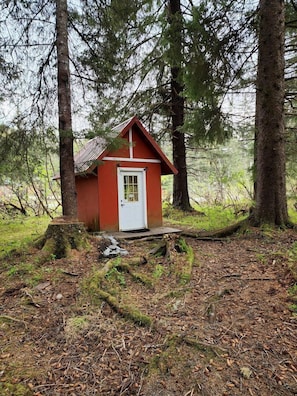 Una cabaña pequeña y roja con una puerta blanca, rodeada de árboles y suelo forestal.