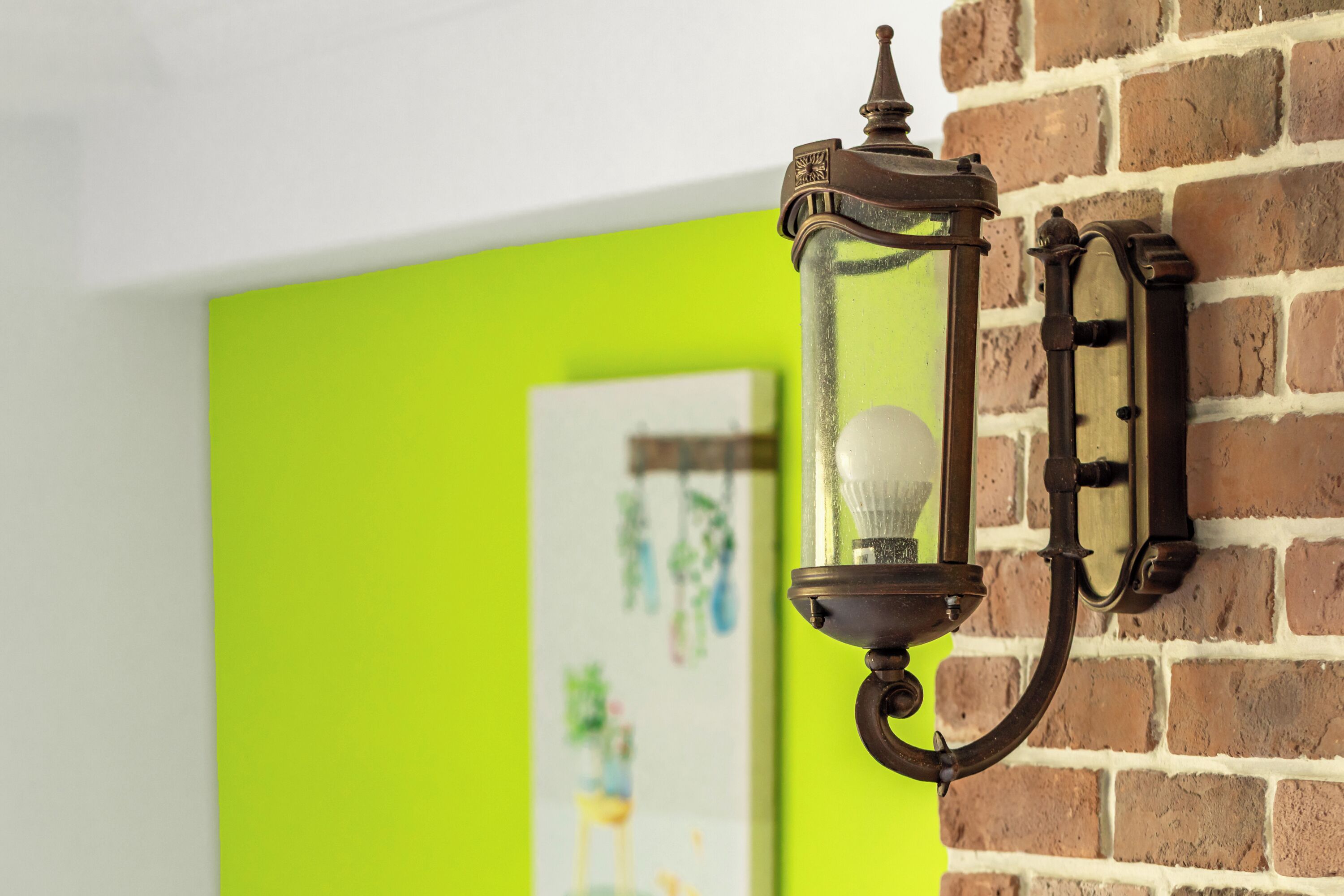A wall-mounted light fixture with a glass enclosure and a bulb, set against a brick wall with a bright green painted section.