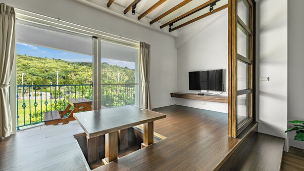 A modern room with a wooden floor, a dining table, a TV mounted on the wall, and a balcony with a view of greenery. - Ghana Inn (Hengchun)