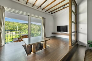 A modern room with a wooden floor, a dining table, a TV mounted on the wall, and a balcony with a view of greenery. - Ghana Inn (Hengchun)