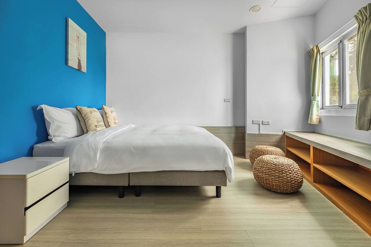 A bedroom with a bed, bedside tables, a window with curtains, a wall art piece, and a woven pouf.
