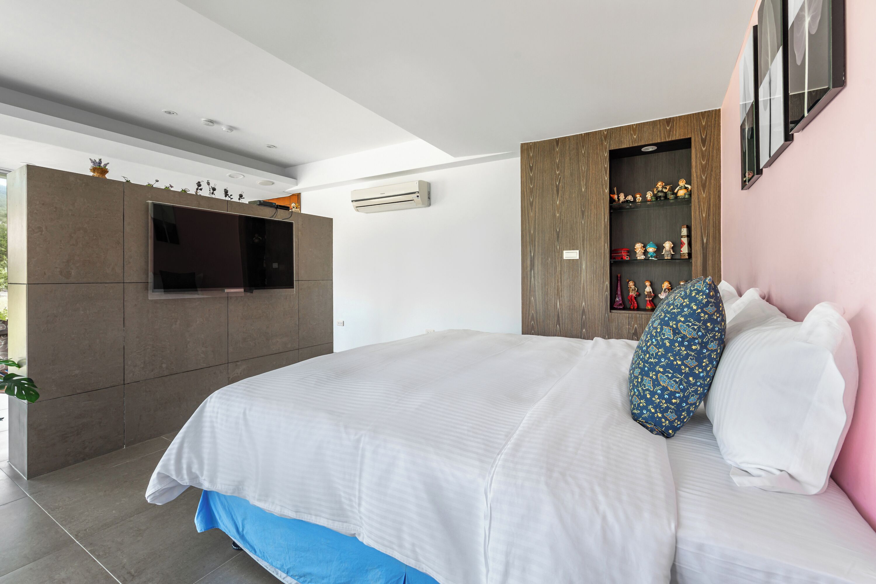 Un dormitorio moderno con una cama grande, un televisor integrado y una estantería de madera con objetos decorativos.
