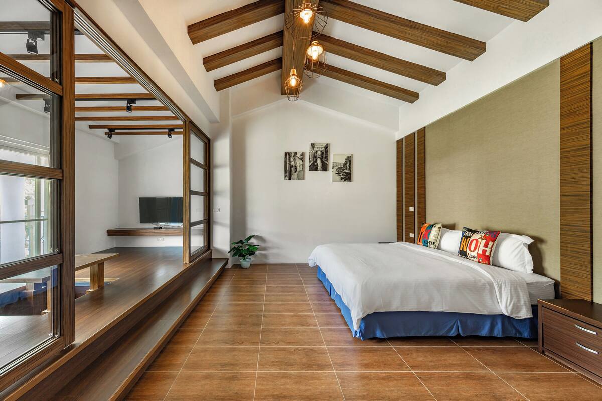 A bedroom with a large bed, wooden beams, a TV, and a window overlooking the room.