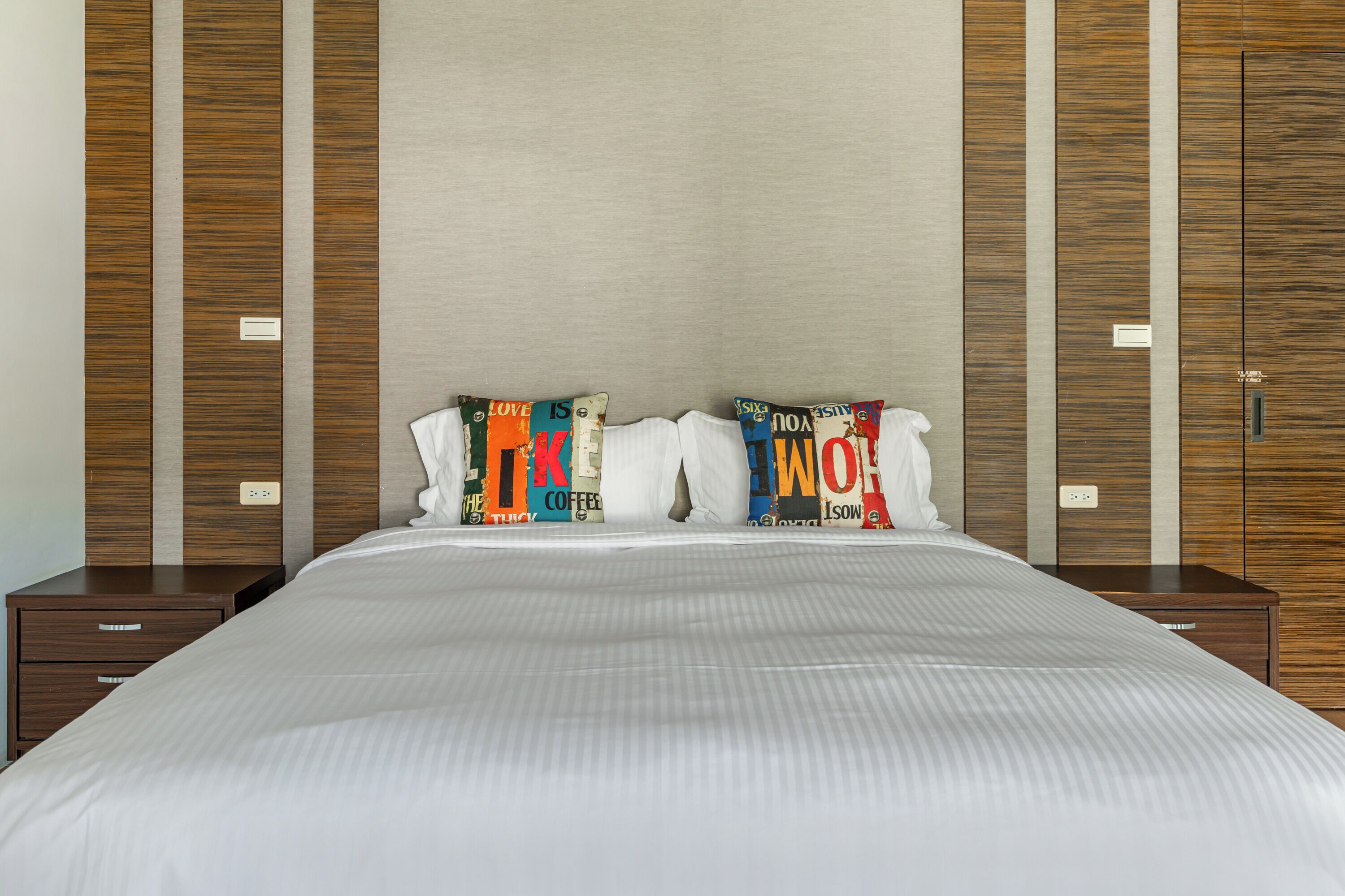 A neatly made bed with a white comforter and two pillows with colorful text. There are wooden panels on the walls and a small wooden nightstand beside the bed.
