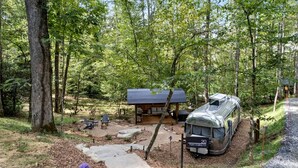 En campingplats omgiven av skog, med ett takförsett område för picknick, en husvagn och en stenlagd stig som leder till ingången.