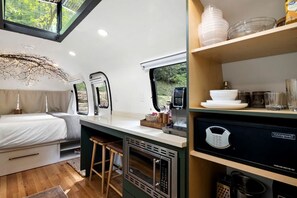A compact RV interior with a kitchenette, a bed, and a window with a view of greenery. - Glamping Getaway in Pisgah Forest in this Lovely Airstream Rental in North Carolina (Pisgah Forest)