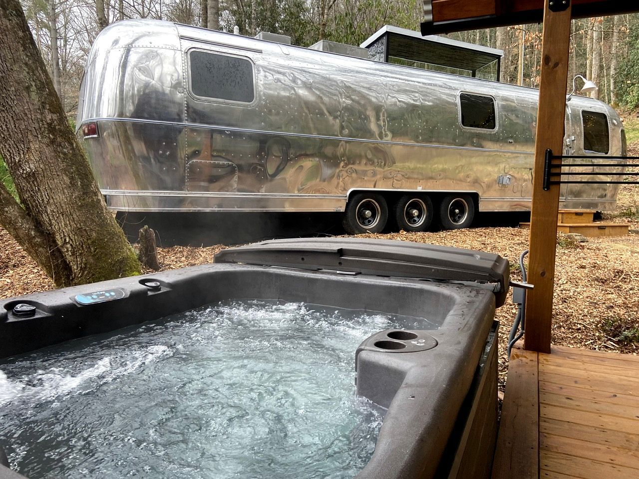 A hot tub with a wooden deck, a silver RV, and a tree in the background.