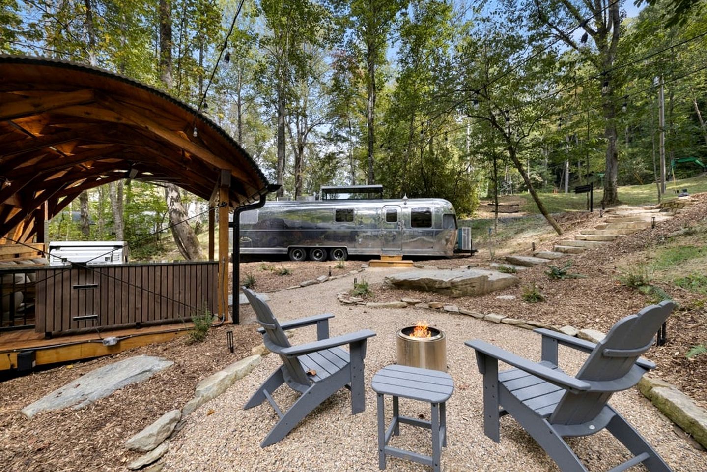 A covered outdoor seating area with a fire pit, a trailer, and a forested backdrop.