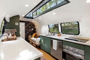 A compact RV kitchen with a central island, a dining area with a sofa, and a window providing a view of the outdoors. - Glamping Getaway in Pisgah Forest in this Lovely Airstream Rental in North Carolina (Pisgah Forest)