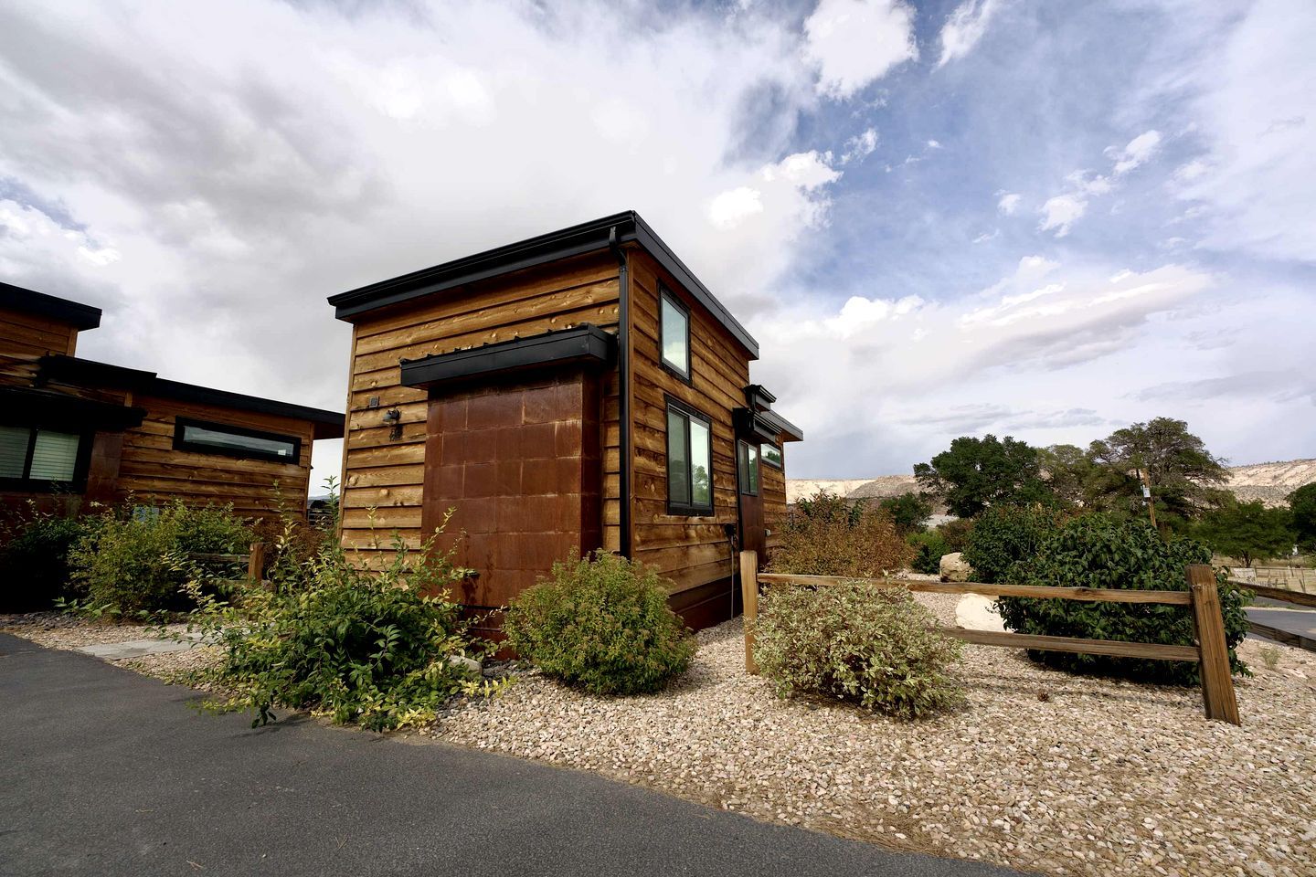 A modern wooden cabin with a large garage door, surrounded by gravel and greenery.