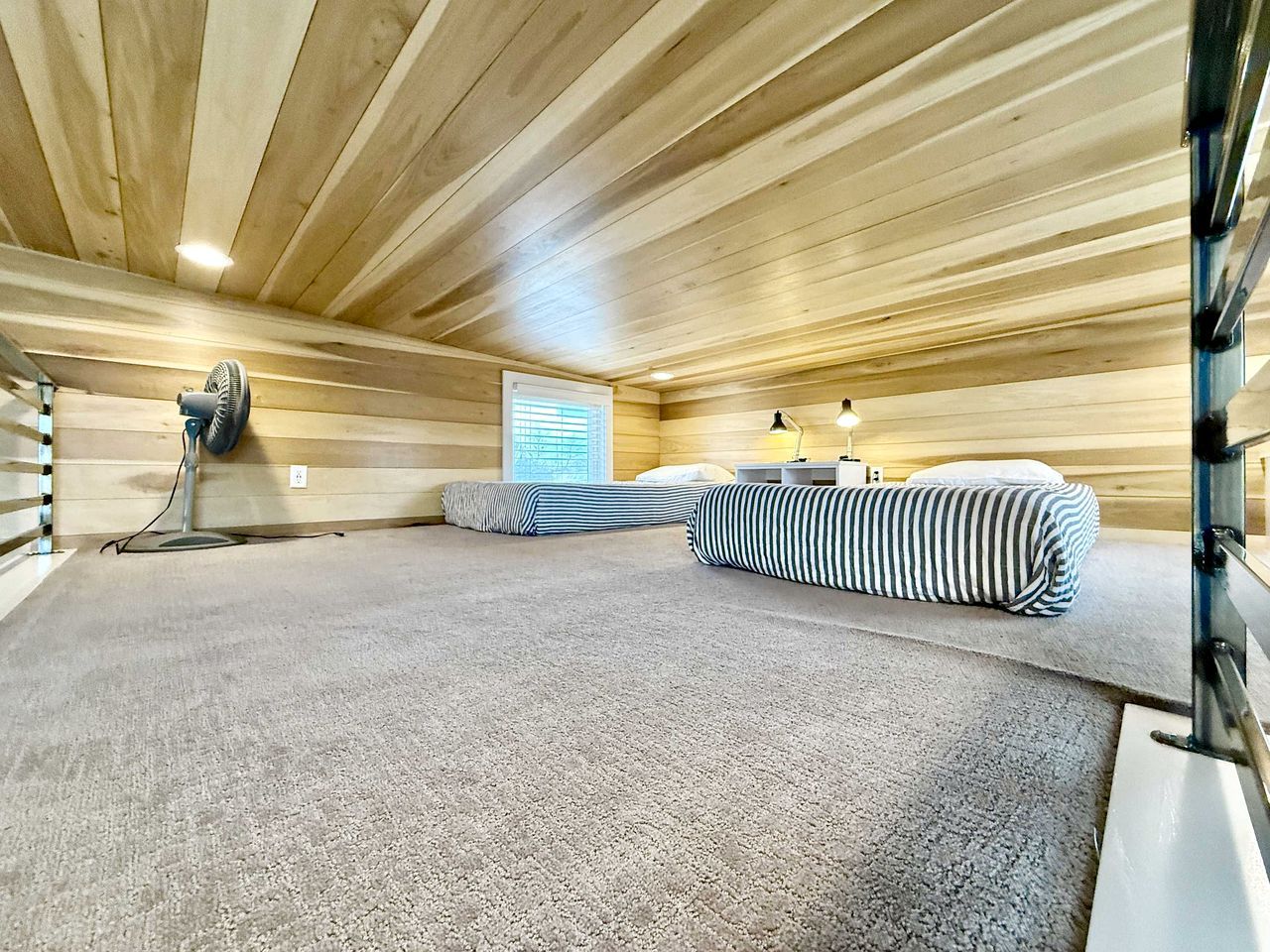 Une pièce dont le sol est recouvert d’une moquette, avec un lit aux draps à rayures, un ventilateur sur pied, une fenêtre avec des stores et un radiateur fixé au mur.