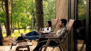 Een houten terras met twee personen zittend op rieten stoelen, met uitzicht op een bebost gebied.