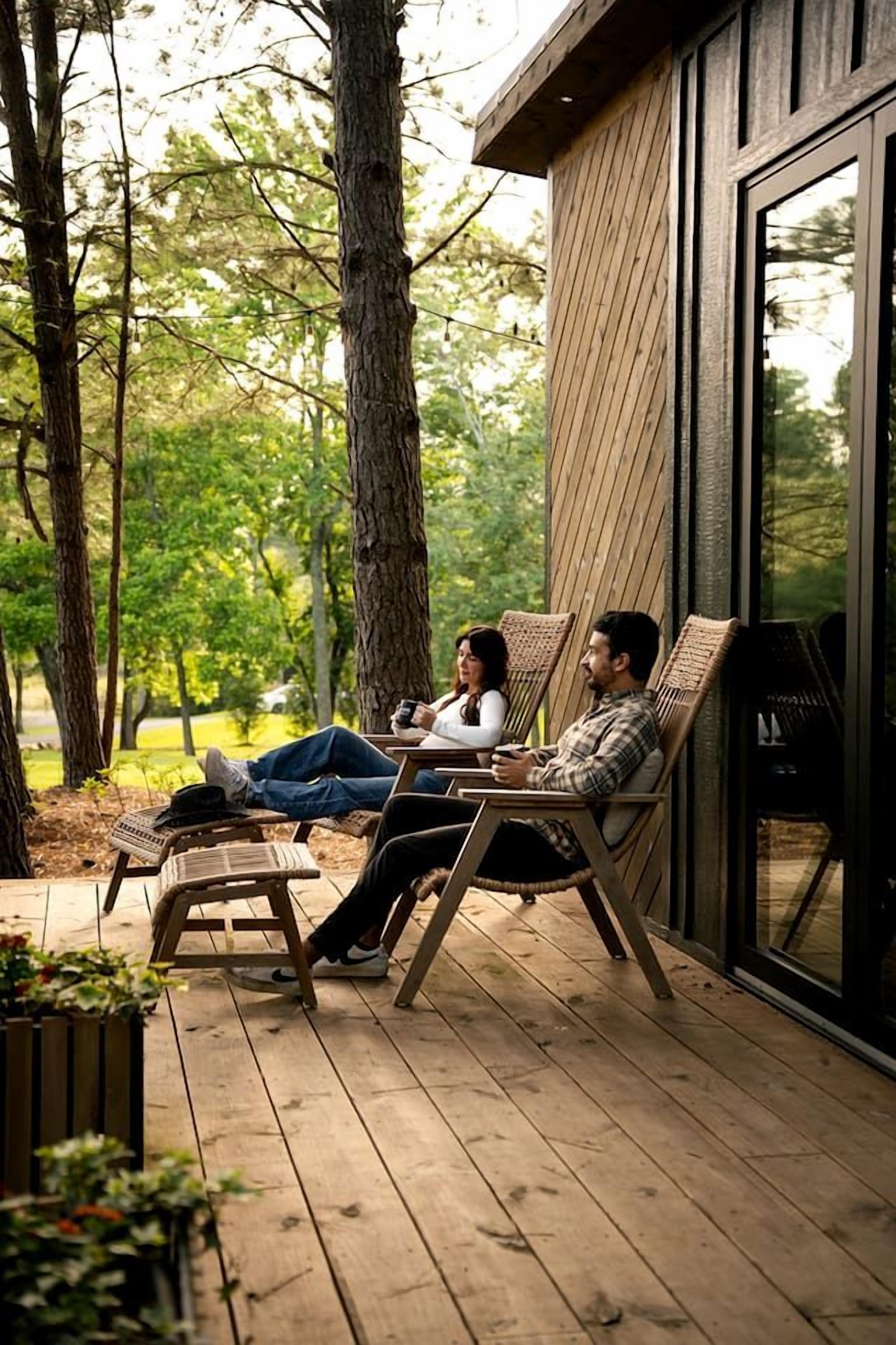 A wooden deck with two people seated on wicker chairs, overlooking a forested area.