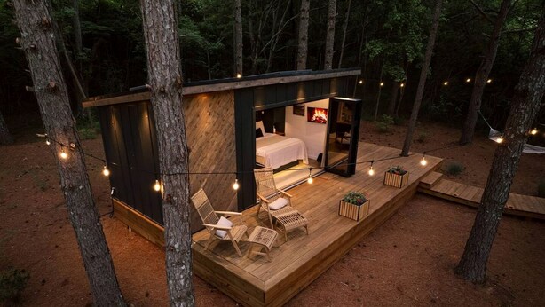 Una cabaña con una cama, dos sillas y una mesita sobre una terraza de madera rodeada de árboles.