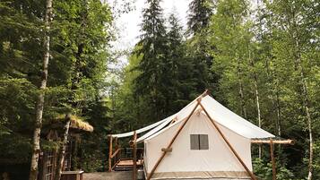 A forest campsite with a large canvas tent, wooden structures, and a gravel path.