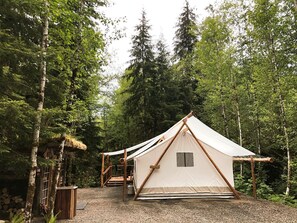 A forest campsite with a large canvas tent, wooden structures, and a gravel path. - Rustic and Spacious Safari Tent in Forks, Washington, Perfect for a Glamping Getaway to the Pacific Northwest (Forks)