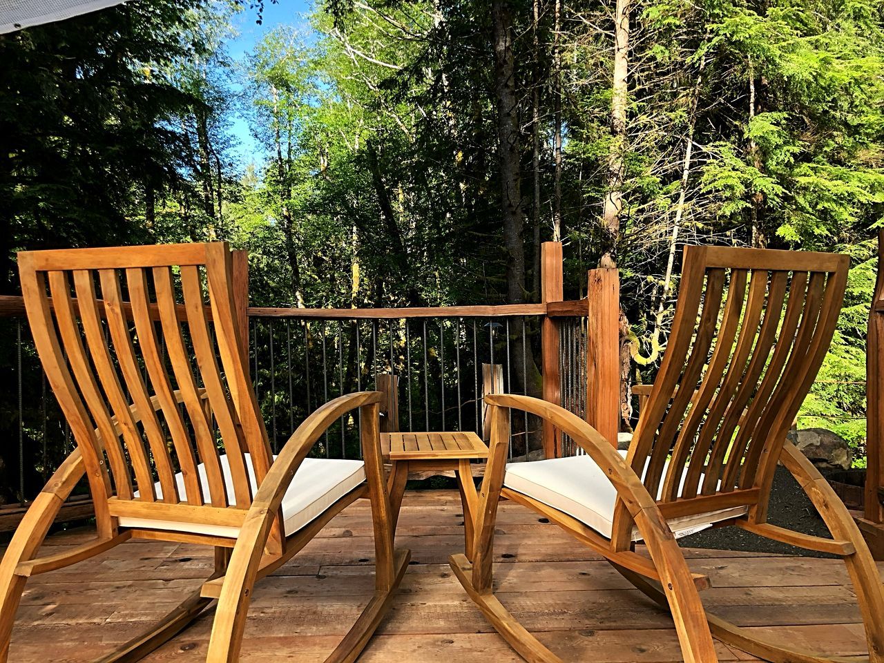 Two wooden Adirondack chairs with cushions on a wooden deck, surrounded by trees.