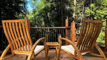 Two wooden Adirondack chairs with cushions on a wooden deck, surrounded by trees.