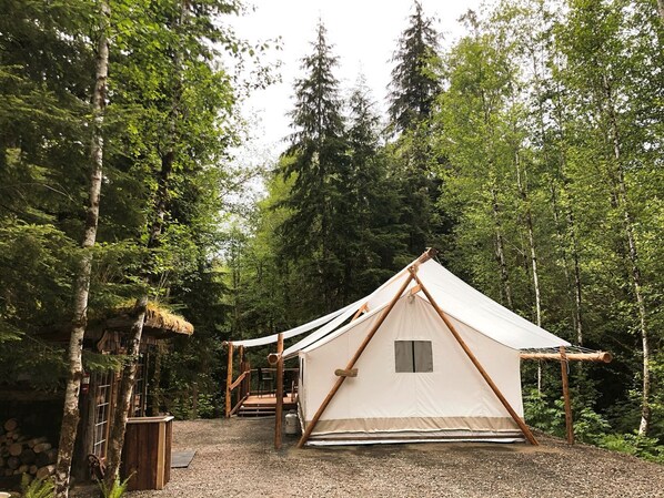 Un terrain de camping situé dans une forêt, avec une grande tente en toile, des structures en bois et un sentier recouvert de gravier.