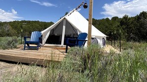 A tent with a wooden deck, two blue chairs, and a wooden post with a hanging object.