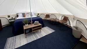 A tented room with a bed, a small table with a plant, a chair, and a waste bin.